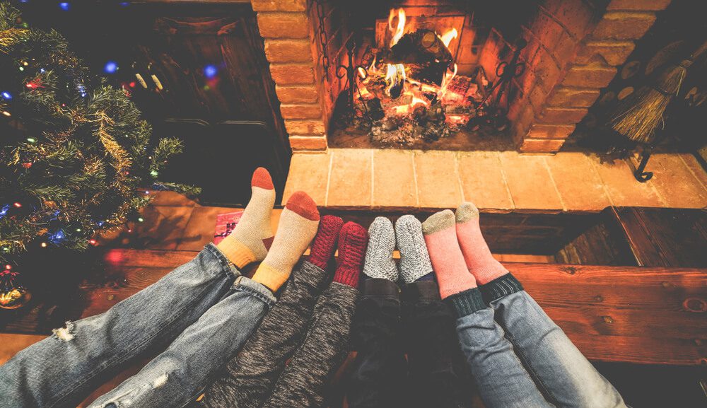 Four legs with thick socks pointed towards a fireplace, next to a christmas tree.